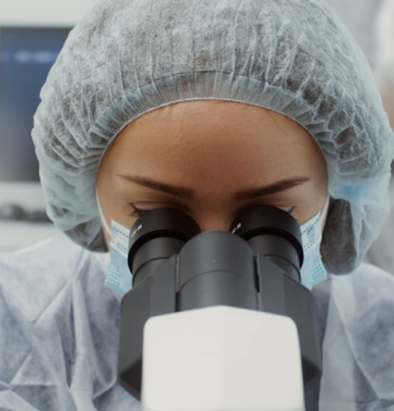 A woman wearing protective clothing and a mask looks through a microscope.